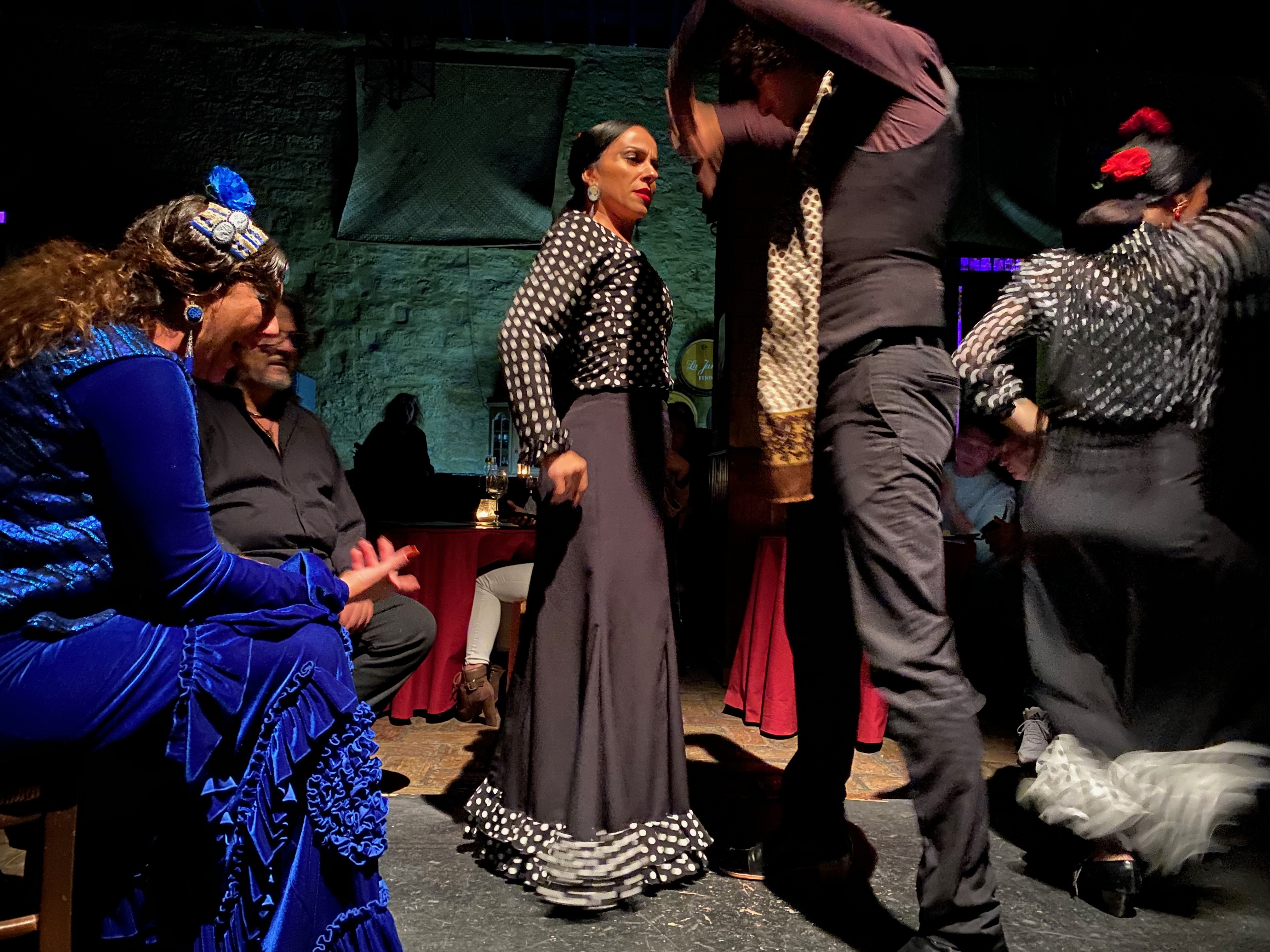 A flamenco tablao in Jerez. Five performers mid-show in an intimate stone-walled room, the cuadro at the edges, the dancer turned toward the male dancer's stamp.